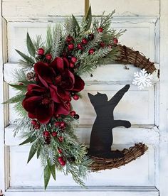 a christmas wreath with red flowers and greenery hanging on the front door, next to a metal cat ornament