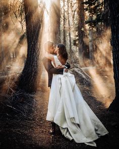 a bride and groom kissing in the woods on their wedding day with sunlight streaming through the trees