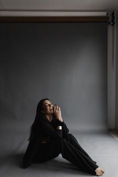 a woman sitting on the floor in front of a gray wall and holding her hands up to her mouth