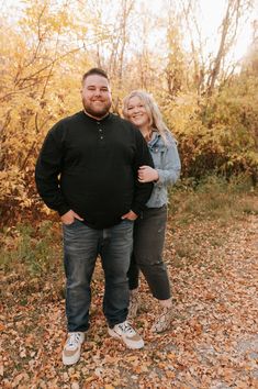 a man and woman standing next to each other in front of trees with leaves on the ground