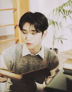 a young man reading a book while sitting at a desk in front of a plant