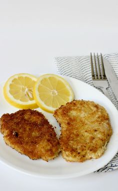 two pieces of fried chicken on a plate with lemon wedges and a fork next to it