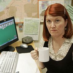 a woman sitting at a desk holding a coffee cup in front of a computer monitor