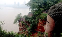 a large buddha statue sitting on top of a cliff next to the ocean with trees growing out of it