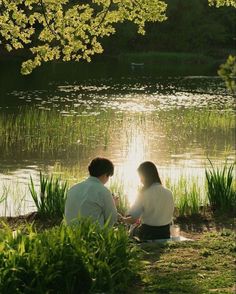 two people sitting on the ground near water and grass, with trees in the background