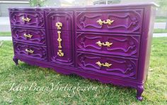 an old dresser painted purple with gold handles and knobs on the drawers is sitting in grass