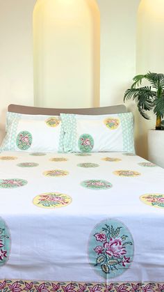 a bed with white sheets and colorful floral designs on the comforter, along with two potted plants