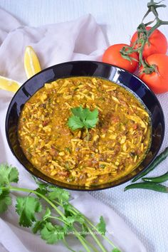 a black bowl filled with yellow curry next to tomatoes and cilantro on a white cloth