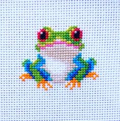 a cross stitch frog sitting on top of a piece of white paper with red eyes