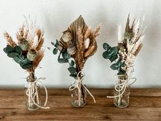 three vases with dried flowers and leaves in them on a wooden table next to a wall