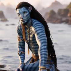 a man dressed in blue and white sitting on rocks near the ocean with long hair