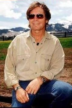 a man wearing sunglasses sitting on a rail in front of a fence with mountains in the background