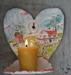 a candle is lit in front of a heart shaped plate with houses painted on it