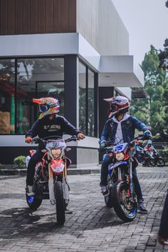 two people riding motorcycles on a brick road in front of a building with large windows