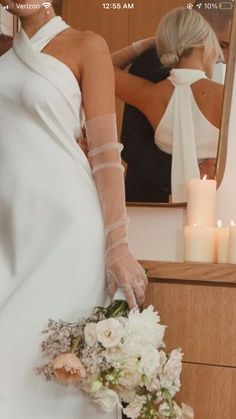 a woman in a white dress is holding a bouquet and looking at herself in the mirror