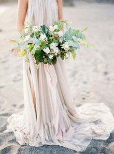 a woman in a white dress holding a bouquet of flowers on the beach with an instagram page below