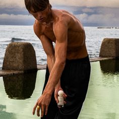 a shirtless man holding an object in his hand near the water's edge