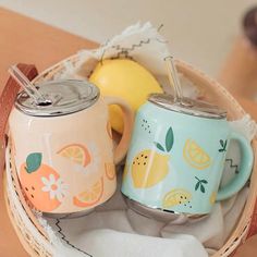 two coffee mugs sitting in a basket with lemons and an apple on the side