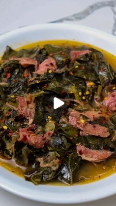 a white bowl filled with greens and meat on top of a table next to a fork