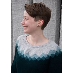 a woman with short hair wearing a green and white sweater smiles at something while standing in front of a wall