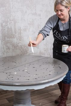 a woman in an apron is painting a table with white paint on it and holding a brush