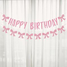 a pink happy birthday banner hanging in front of a window with sheer curtains and white drapes