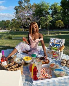 a woman sitting at a picnic table with food and drinks in front of her on the grass