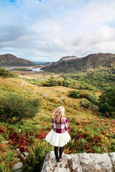 a woman standing on top of a rock in the middle of a field with mountains behind her