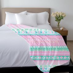 a bed with white sheets and pink and green blankets on it, next to a vase with flowers