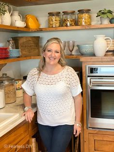 a woman is standing in the kitchen next to an oven and shelves with jars on them