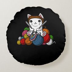 a black round pillow with a cartoon cat sitting on top of balls of yarn and knitting needles