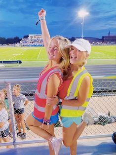 two young women standing next to each other in front of a fence at a baseball field