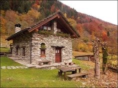 an old stone house with a wooden bench in front of it and autumn foliage on the hillside behind