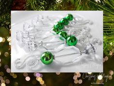 three green glass ornaments hanging from a christmas ornament on a pine tree with lights in the background