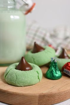 green cookies with chocolate on top and candy in the middle, sitting on a cutting board next to a jar of milk