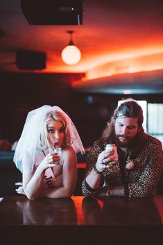 two people sitting at a table with drinks in their hands and one person wearing a veil