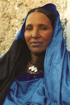 a woman wearing a blue shawl sitting next to a stone wall and looking at the camera