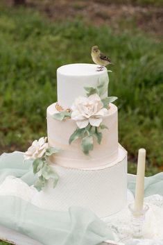 a white wedding cake with flowers and birds on top sitting on a table outside in the grass