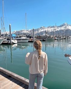 a woman standing on a dock looking at boats in the water and snow covered mountains