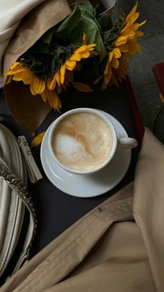 a cup of coffee sitting on top of a table next to a vase with sunflowers