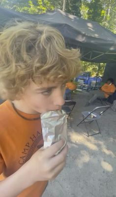 a young boy is eating something out of a tin foil wrapper in front of an outdoor tent