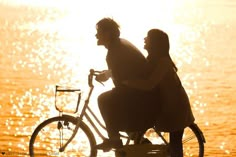 a man and woman riding on the back of a bike next to water at sunset