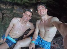 two men in swim trunks sitting next to each other on the ground near water and rocks