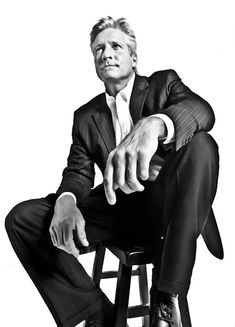 a man in a suit and tie sitting on a stool with his hand out to the side