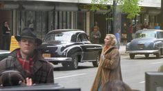 a man and woman walking down the street in front of old cars on a busy city street