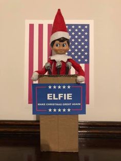 an elf is standing behind a sign that says elfie make christmas great again