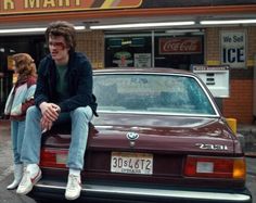 two people sitting on the hood of a car in front of a store with an ice cream parlor