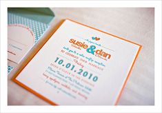 an orange and white wedding card on top of a table next to a green envelope