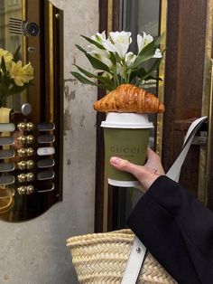 a woman holding a cup with a croissant on top of it and a straw bag in front of her