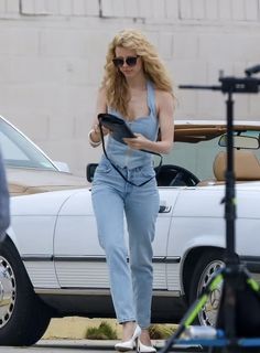 a woman in overalls and heels is looking at something on her tablet while standing next to a car